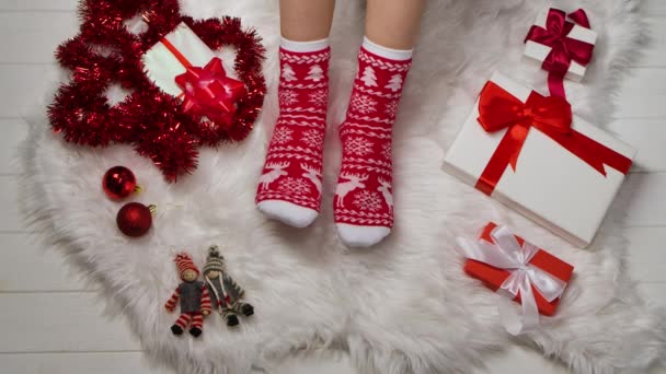 Patas femeninas en calcetines cálidos con adornos rojos blancos sobre un fondo de piel de piel, oropel y cajas con regalos relajantes en casa. Vacaciones de invierno Navidad y Año Nuevo concepto. De cerca. Movimiento lento. — Vídeos de Stock