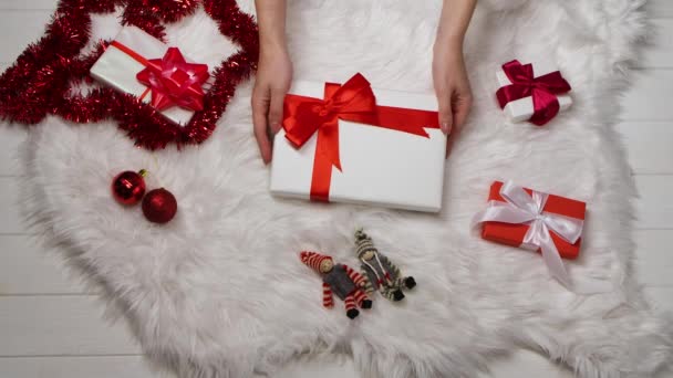 A női kezek egy ajándékdobozt helyeznek egy fehér szőrme bőrre, egy szétszórt vörös rózsa köré, és dekorációkat a karácsonyfához. Téli ünnepek karácsony és újév. Közelről. Lassú mozgás.. — Stock videók