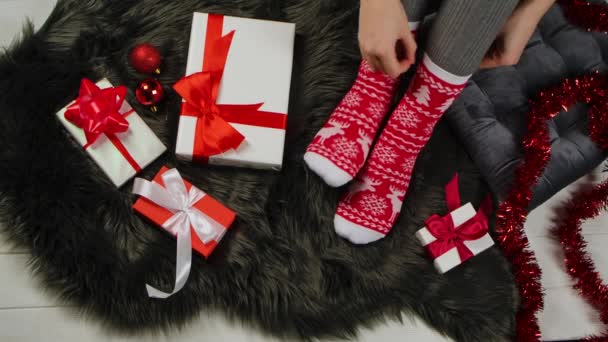 Женщина в рождественских теплых носках с красными и белыми орнаментами сидит на серой меховой коже. Подарки, украшения и мишура разбросаны повсюду. Праздник. С Рождеством и Новым годом. Закрывай. Медленное движение. — стоковое видео