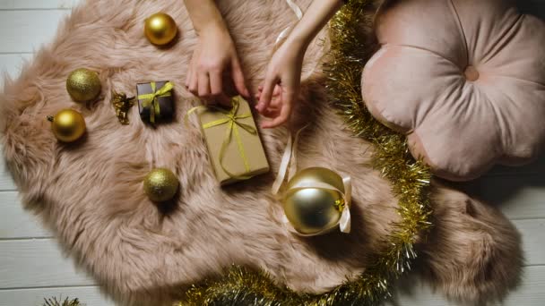 As mãos de mulheres endireitam um arco em uma caixa de presente que está em uma pele de pele em volta de ouropel e decorações de Natal coloridas de ouro. Fundo festivo. Preparando-se para Boas Festas de Inverno. Fecha. Movimento lento. — Vídeo de Stock