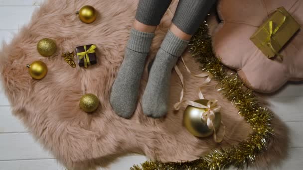 Femme en chaussettes grises s'assoit sur une peau de fourrure autour de boîtes avec des cadeaux et des décorations de Noël de couleur or. Le concept de vacances agréables et heureuses à la maison. Joyeux Noël et nouvel an. Ferme là. Mouvement lent. — Video