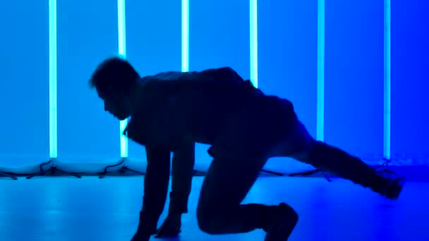 Karanlık bir stüdyoda açık mavi neon ışıkların önünde dans eden şık bir genç adamın silueti. Kapatın. Yavaş çekim. — Stok video