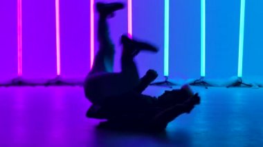Experienced dancer performs complex break dance elements, rotates on the floor and does handstand. Silhouette of a man against the background of bright multicolored neon lights. Close up. Slow motion.