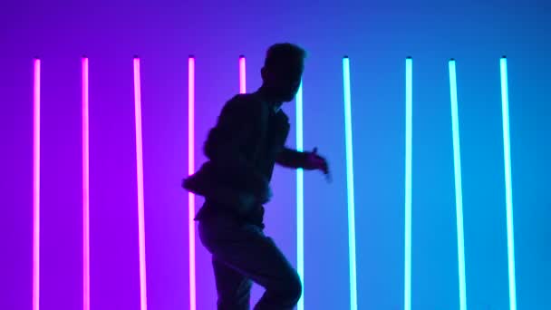 De camera beweegt zich naar een stijlvolle man dynamisch dansend breakdance en hip hop tegen de achtergrond van heldere veelkleurige neon lichten. Silhouet van mannelijke danser van dichtbij. Langzame beweging. — Stockvideo