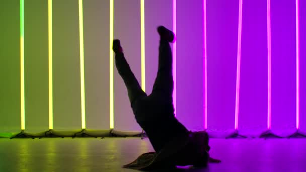 Profesjonalny breaktancer wykonuje skomplikowane obroty na podłodze. Sylwetka mężczyzny tańczącego na tle jasnych neonów w studiu o zmierzchu. Zamknij drzwi. Zwolniony ruch. — Wideo stockowe