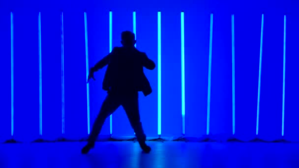 Silhouette of a young man in stylish youth clothes dancing breakdance street dance against the backdrop of bright blue neon lights in a dark studio. Slow motion. — Stock Video