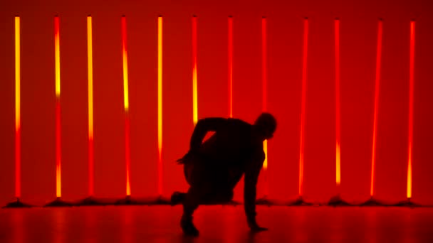 Stylish male break dance dancer performs a spin on the floor and handstands in a dark studio against a background of red and yellow neon tubes. Silhouette of man performing complex tricks. Slow motion — Stock Video