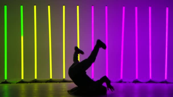 El breakdancer profesional realiza giros complejos en el suelo. Silueta de un hombre bailando sobre el telón de fondo de brillantes luces de neón en un estudio crepuscular. Movimiento lento. — Vídeos de Stock