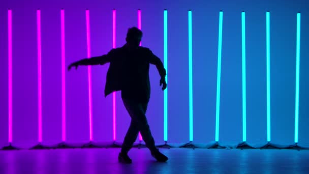 Young stylish man jumping and dancing breakdance in street style. A dancer practices his skills against the backdrop of bright neon lights in a dark studio. Silhouette. Slow motion. — Stock Video