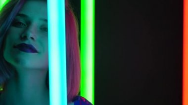 Pembe kısa saçlı, şık bir sarışının portresi parlak neon ışıklı, karanlık bir stüdyo arka planına karşı poz veriyor. Kapatın. Yavaş çekim.