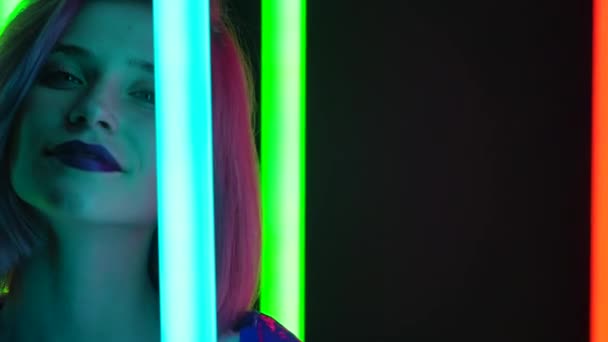 Retrato de uma jovem loira sexual elegante com cabelo curto rosa posa contra um fundo de estúdio escuro com luzes de néon multicoloridas brilhantes. Fecha. Movimento lento. — Vídeo de Stock
