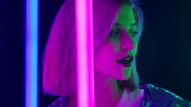 Pembe kısa saçlı, şık bir sarışının portresi parlak neon ışıklı, karanlık bir stüdyo arka planına karşı poz veriyor. Kapatın. Yavaş çekim.