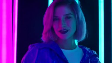 Pembe kısa saçlı, şık bir sarışının portresi parlak neon ışıklı, karanlık bir stüdyo arka planına karşı poz veriyor. Kapatın. Yavaş çekim.