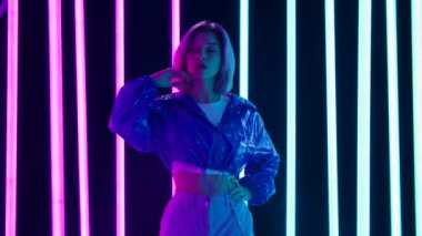 Pembe kısa saçlı, şık bir sarışının portresi parlak neon ışıklı, karanlık bir stüdyo arka planına karşı poz veriyor. Yavaş çekim.