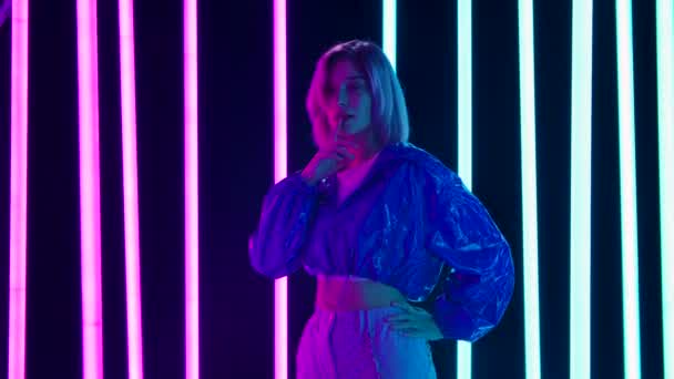 Portrait d'une jeune blonde sexuelle élégante aux cheveux courts roses pose sur un fond de studio sombre avec des lumières au néon multicolores lumineuses. Mouvement lent. — Video