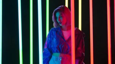 Pembe kısa saçlı, şık bir sarışının portresi parlak neon ışıklı, karanlık bir stüdyo arka planına karşı poz veriyor. Yavaş çekim.