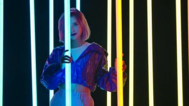 Pembe kısa saçlı, şık bir sarışının portresi parlak neon ışıklı, karanlık bir stüdyo arka planına karşı poz veriyor. Yavaş çekim.