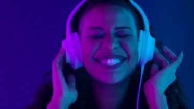 Kameraya bakıp büyük beyaz kulaklıklarla müzik dinlemekten hoşlanan güzel bir Afrikalı kadının portresi. Kapalı yüz, mor ve mavi neon ışıklarla aydınlatılmış. Yavaş çekim.