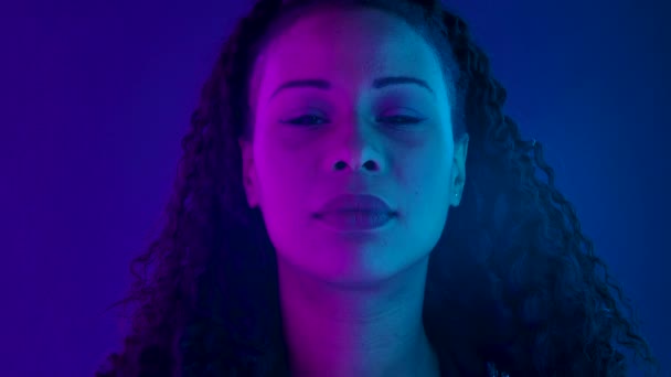 Portret van een jonge mooie Afro-Amerikaanse vrouw die naar de camera kijkt. Close-up gezicht verlicht met paarse en blauwe neon lichten. Langzame beweging. — Stockvideo