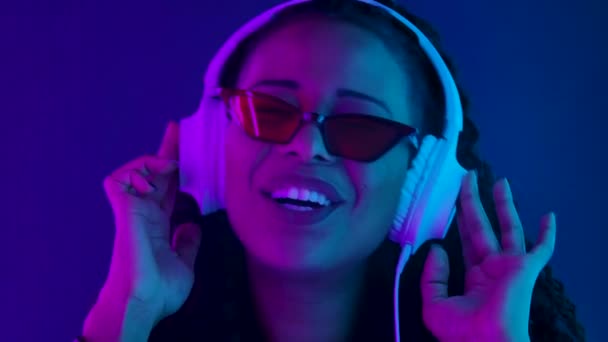 Retrato de uma jovem adorável mulher afro-americana gosta de ouvir música em grandes fones de ouvido brancos.Close-up rosto iluminado com luzes de néon roxo e azul. Movimento lento. — Vídeo de Stock