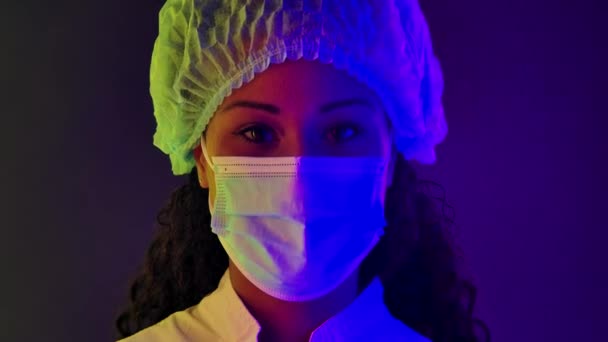 Porträtt av en ung vacker kvinna afroamerikan i skyddskläder och en medicinsk mask tittar på kameran. Närbild ansikte upplyst med flerfärgade neonljus. Långsamma rörelser. — Stockvideo