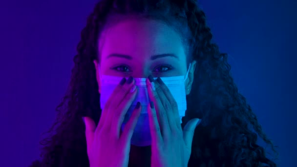 Retrato de una joven y encantadora mujer afroamericana mirando a la cámara se pone y luego se quita la máscara médica. Cara cercana iluminada con luces de neón púrpura y azul. Movimiento lento. — Vídeos de Stock