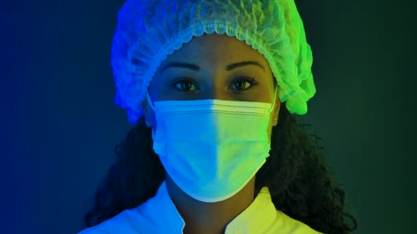 Porträtt av en ung vacker kvinnlig afroamerikan i skyddskläder, tar bort den medicinska masken med lättnad. Närbild ansikte upplyst med flerfärgade neonljus. Långsamma rörelser. — Stockvideo