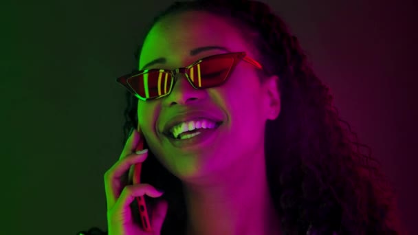 スマートフォンで話すスタイリッシュなサングラスで若い素敵な女性アフリカ系アメリカ人の肖像画。多色のネオンライトで照らされた顔を閉じます。スローモーション. — ストック動画
