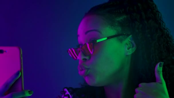 Porträtt av en ung vacker kvinna afroamerikan i snygga solglasögon, känslomässigt talar via videosamtal på sin smartphone. Närbild ansikte upplyst med flerfärgade neonljus. Långsamma rörelser. — Stockvideo
