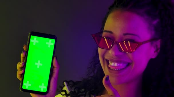 Porträt einer jungen hübschen Afroamerikanerin zeigt mit dem Finger auf das Smartphone mit grünem Bildschirmschlüssel und zeigt eine Daumen-hoch-Geste. Aus nächster Nähe. Zeitlupe. — Stockvideo