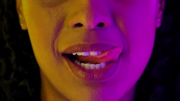 Aantrekkelijke Afro-Amerikaanse lippen en mond. Jonge vrouw glimlacht, likt en bijt op zijn lippen. Close-up macro portret verlicht door fel neon licht. Langzame beweging. — Stockvideo