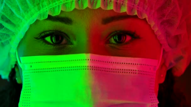 Potret seorang wanita muda Afrika Amerika yang cantik dengan topi pelindung dan topeng medis melihat ke kamera. Tutup potret makro yang diterangi dengan lampu neon warna-warni. Gerakan lambat. — Stok Video