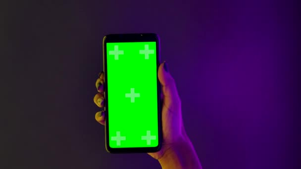 Mujer afroamericana sostiene un teléfono inteligente en su mano y toca la tecla croma pantalla verde. Cierre el teléfono en posición vertical sobre un fondo de luces de neón multicolores brillantes. Movimiento lento. — Vídeos de Stock