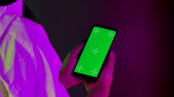 Vue de derrière une femme afro-américaine tenant un smartphone avec une clé chromatique à écran vert à la main. Gros plan d'un téléphone sur un fond de lumières au néon multicolores lumineuses. Mouvement lent. — Video