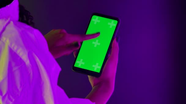 Vedeți din spatele unei femei afro-americane care deține un smartphone în mână și atinge un ecran verde cu o cheie de cromă. Închide telefonul pe un fundal de lumini luminoase de neon. Slow motion . — Videoclip de stoc