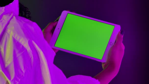 Blick von hinten auf eine afroamerikanische Frau, die ein weißes Tablet in der Hand hält und einen grünen Screen-Chroma-Schlüssel berührt. Großaufnahme eines Gadgets vor einem Hintergrund heller Neonlichter. Zeitlupe. — Stockvideo