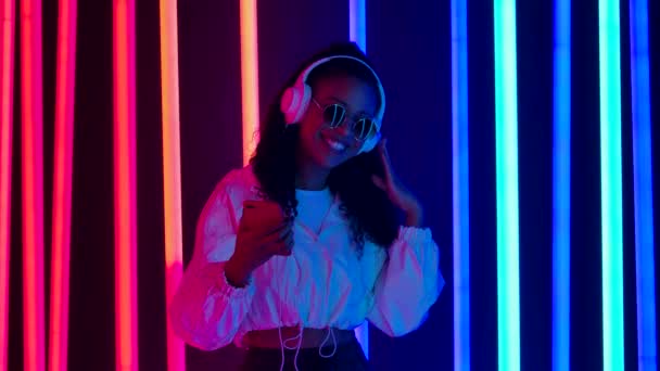 Retrato de una joven encantadora mujer afroamericana está bailando y disfrutando de la música desde el teléfono inteligente en grandes auriculares blancos. Movimiento lento. — Vídeos de Stock