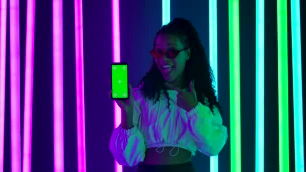 Retrato de una joven y encantadora mujer afroamericana con gafas de sol elegantes apunta con el dedo a un teléfono inteligente con una tecla de croma de pantalla verde. Movimiento lento. — Vídeos de Stock