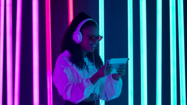 Retrato de una joven encantadora mujer afroamericana juega un juego en una tableta con grandes auriculares blancos y se regocija en la victoria. Movimiento lento. — Vídeos de Stock