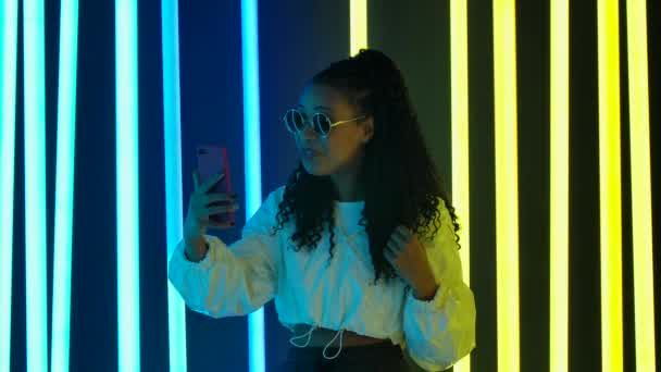 Porträt einer jungen hübschen Afroamerikanerin mit stylischer Sonnenbrille, die per Videoanruf auf ihrem Smartphone spricht, Kaugummi kaut und eine Blase pustet. Zeitlupe. — Stockvideo