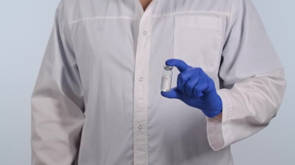 De arts in een wit medisch pak en beschermende handschoenen houdt de injectieflacon met coronavirusvaccin in zijn hand. De mens adviseert het vaccin te gebruiken voor immunisatie tegen het COVID-virus. Sluit maar af. Langzame beweging. — Stockvideo