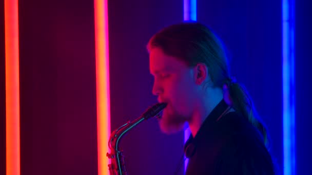 Vista lateral de un hombre joven y elegante con el pelo largo tocando el saxofón magistralmente. El músico realiza un concierto en vivo en un estudio oscuro con el telón de fondo de brillantes luces de neón. De cerca. Movimiento lento. — Vídeo de stock