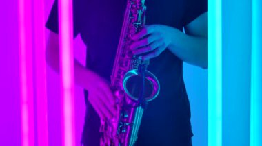 Erkek eller saksafonun anahtarlarını parmaklıyor. Bir saksafoncu, karanlık bir stüdyoda parlak neon ışıkların önünde konser verir. Kapatın. Yavaş çekim.