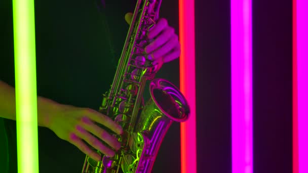 Gedetailleerde beelden van de mannelijke handen van een muzikant die een klassieke melodie uitvoert op een saxofoon. Geschoten in een donkere studio met heldere neon buizen op de achtergrond. Sluit maar af. Langzame beweging. — Stockvideo