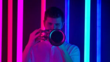 Trompet çalan bir müzisyenin canlı performansı. Beyaz gömlekli bir adam, karanlık bir stüdyoda parlak neon ışıkların arka planına karşı caz melodisi çalar. Kapatın. Yavaş çekim.