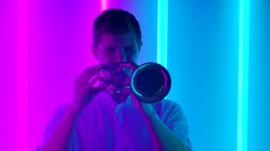 Profesyonel bir trompetçiden caz solo performansı. Beyaz gömlekli bir adam, karanlık bir stüdyoda, parlak neon ışıkların arka planına karşı gösteri yapıyor. Müzikal şov. Kapatın. Yavaş çekim.