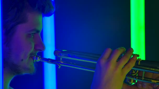 Vista lateral de un joven trompetista profesional que actúa en un espectáculo nocturno entre luces de neón brillantes. Un hombre toca un instrumento de bronce en vivo. De cerca. Movimiento lento. — Vídeos de Stock