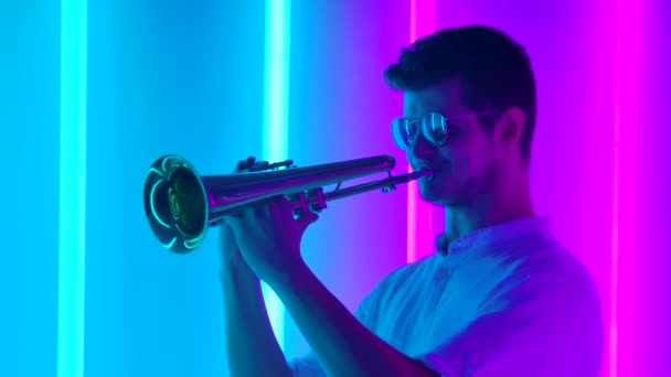 Young stylish man in sunglasses plays the trumpet enthusiastically. Music party in retro style with bright colored neon lights. Close up. Slow motion. — Stock Video