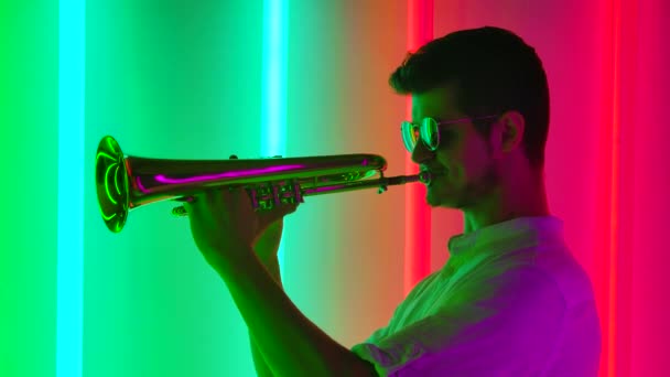 Joven hombre con estilo en gafas de sol toca la trompeta con entusiasmo. Fiesta de música en estilo retro con luces de neón de colores brillantes. De cerca. Movimiento lento. — Vídeo de stock