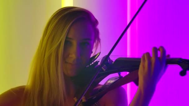 Ung attraktiv blondin spelar fiol bland de ljusa neonljus. En kvinna vidrör stråkarna på bågarna när hon spelar klassisk musik. Närbild. Långsamma rörelser. — Stockvideo